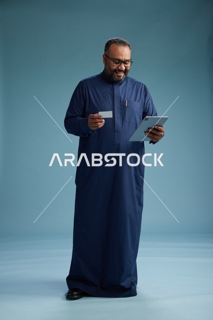 A portrait of a Saudi man wearing a blue thobe holding a blank white card in one hand while using a tablet, electronic payment via a bank card, full body, blue background.