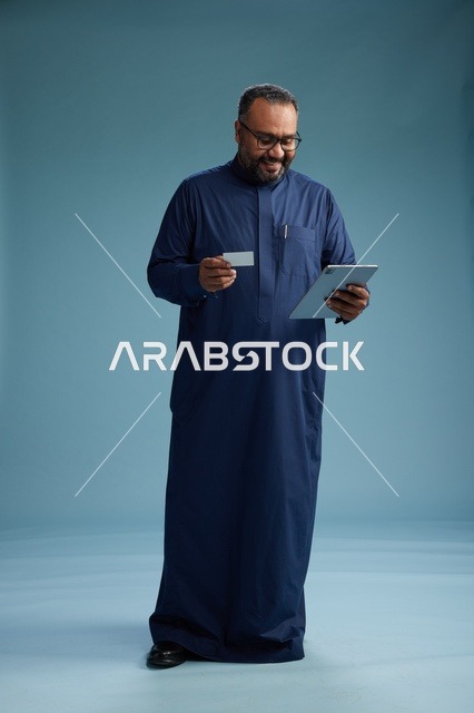 A portrait of a Saudi man wearing a blue thobe holding a blank white card in one hand while using a tablet, electronic payment via a bank card, full body, blue background.