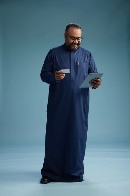 A portrait of a Saudi man wearing a blue thobe holding a blank white card in one hand while using a tablet, electronic payment via a bank card, full body, blue background.