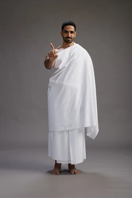 A portrait of an Arab Saudi Gulf man wearing Ihram clothing to perform the rituals of Hajj, worship, and drawing closer to God, Hajj and Umrah in Mecca, full-body, gray background.