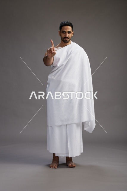 بورتريه لرجل عربي سعودي خليجي يرتدي ملابس الإحرام لأداء مناسك الحج, العباده والتقرب الى الله , الحج والعمره في مكه, الجسم بالطول الكامل , خلفيه رماديه