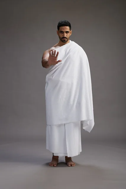 A portrait of an Arab Saudi Gulf man wearing Ihram clothing to perform the rituals of Hajj, worship, and drawing closer to God, Hajj and Umrah in Mecca, full-body, gray background.