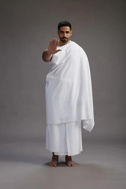 A portrait of an Arab Saudi Gulf man wearing Ihram clothing to perform the rituals of Hajj, worship, and drawing closer to God, Hajj and Umrah in Mecca, full-body, gray background.