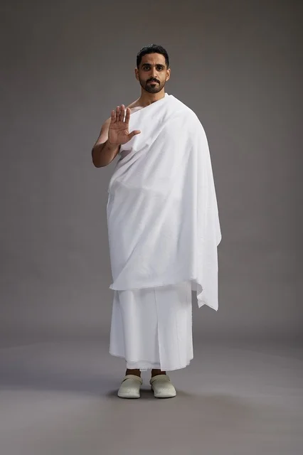 A portrait of an Arab Saudi Gulf man wearing Ihram clothing to perform the rituals of Hajj, worship, and drawing closer to God, Hajj and Umrah in Mecca, full-body, gray background.