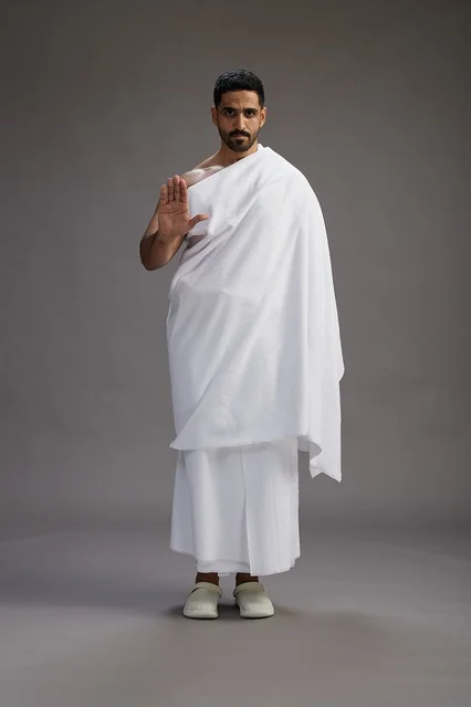 A portrait of an Arab Saudi Gulf man wearing Ihram clothing to perform the rituals of Hajj, worship, and drawing closer to God, Hajj and Umrah in Mecca, full-body, gray background.