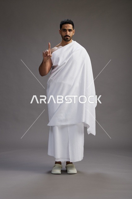 بورتريه لرجل عربي سعودي خليجي يرتدي ملابس الإحرام لأداء مناسك الحج, العباده والتقرب الى الله , الحج والعمره في مكه, الجسم بالطول الكامل , خلفيه رماديه