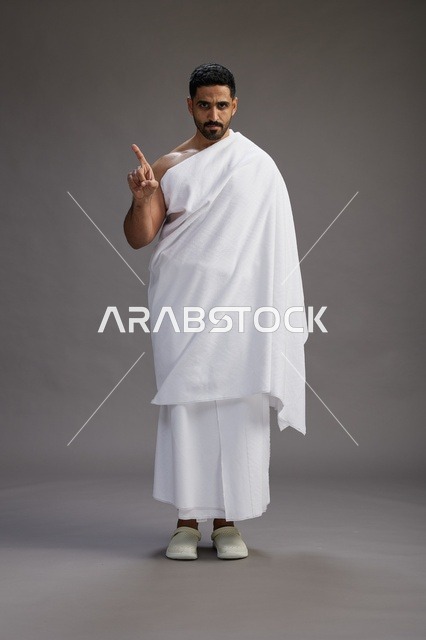 A portrait of an Arab Saudi Gulf man wearing Ihram clothing to perform the rituals of Hajj, worship, and drawing closer to God, Hajj and Umrah in Mecca, full-body, gray background.