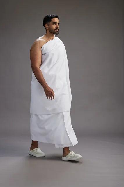 A portrait of an Arab Saudi Gulf man wearing Ihram clothing to perform the rituals of Hajj, worship, and drawing closer to God, Hajj and Umrah in Mecca, full-body, gray background.