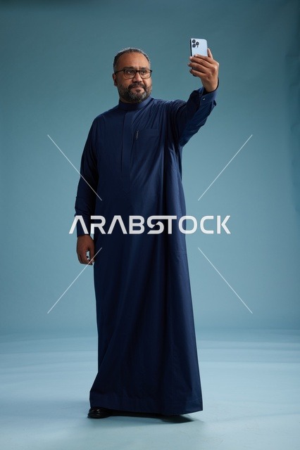 بورتريه لرجل عربي  سعودي  خليجي بثوب أزرق يلتقط سيلفي باستخدام الهاتف المحمول, ايماءات السعاده والضحك, اشارات وحركات متنوعه , الجسم بالطول الكامل, خلفيه زرقاء