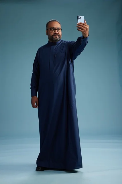A portrait of an Arab Saudi Gulf man in a blue thobe taking a selfie with a mobile phone, expressions of happiness and laughter, various gestures and movements, full-body, blue background.