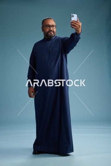 بورتريه لرجل عربي  سعودي  خليجي بثوب أزرق يلتقط سيلفي باستخدام الهاتف المحمول, ايماءات السعاده والضحك, اشارات وحركات متنوعه , الجسم بالطول الكامل, خلفيه زرقاء