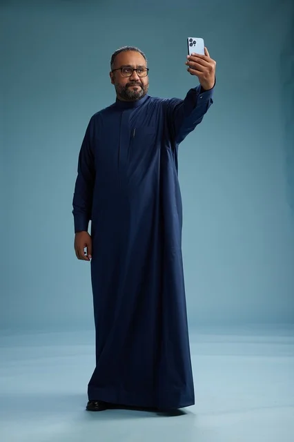 A portrait of an Arab Saudi Gulf man in a blue thobe taking a selfie with a mobile phone, expressions of happiness and laughter, various gestures and movements, full-body, blue background.