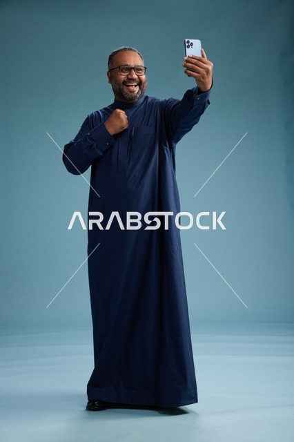 بورتريه لرجل عربي  سعودي  خليجي بثوب أزرق يلتقط سيلفي باستخدام الهاتف المحمول, ايماءات السعاده والضحك, اشارات وحركات متنوعه , الجسم بالطول الكامل, خلفيه زرقاء