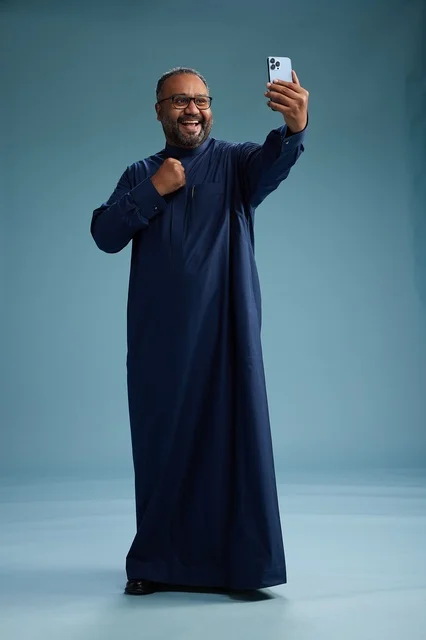 بورتريه لرجل عربي  سعودي  خليجي بثوب أزرق يلتقط سيلفي باستخدام الهاتف المحمول, ايماءات السعاده والضحك, اشارات وحركات متنوعه , الجسم بالطول الكامل, خلفيه زرقاء