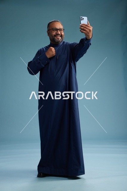 بورتريه لرجل عربي  سعودي  خليجي بثوب أزرق يلتقط سيلفي باستخدام الهاتف المحمول, ايماءات السعاده والضحك, اشارات وحركات متنوعه , الجسم بالطول الكامل, خلفيه زرقاء