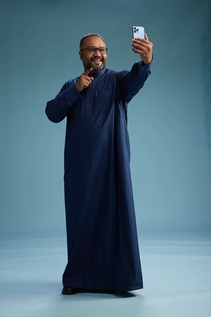 A portrait of an Arab Saudi Gulf man in a blue thobe taking a selfie with a mobile phone, expressions of happiness and laughter, various gestures and movements, full-body, blue background.