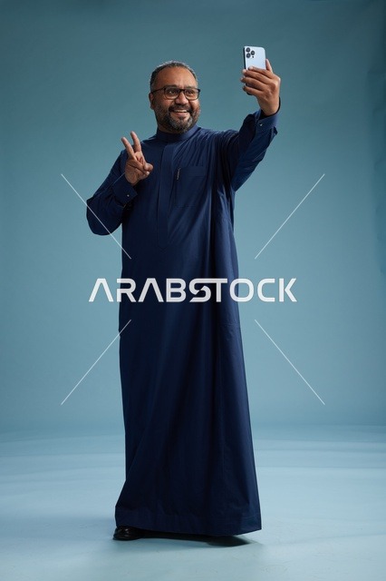 بورتريه لرجل عربي  سعودي  خليجي بثوب أزرق يلتقط سيلفي باستخدام الهاتف المحمول, ايماءات السعاده والضحك, اشارات وحركات متنوعه , الجسم بالطول الكامل, خلفيه زرقاء
