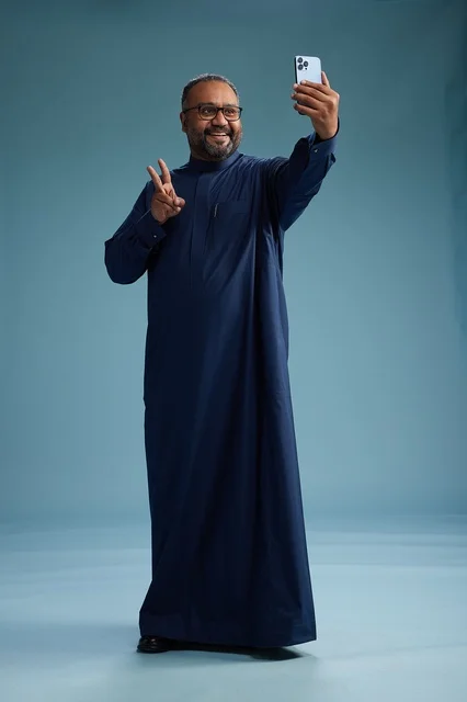 A portrait of an Arab Saudi Gulf man in a blue thobe taking a selfie with a mobile phone, expressions of happiness and laughter, various gestures and movements, full-body, blue background.