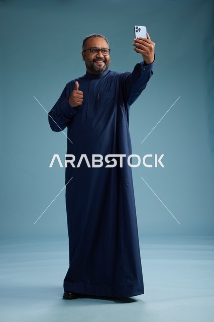 بورتريه لرجل عربي  سعودي  خليجي بثوب أزرق يلتقط سيلفي باستخدام الهاتف المحمول, ايماءات السعاده والضحك, اشارات وحركات متنوعه , الجسم بالطول الكامل, خلفيه زرقاء