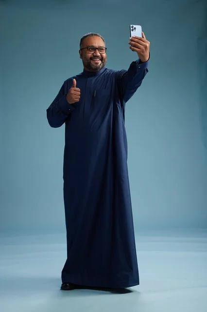 A portrait of an Arab Saudi Gulf man in a blue thobe taking a selfie with a mobile phone, expressions of happiness and laughter, various gestures and movements, full-body, blue background.