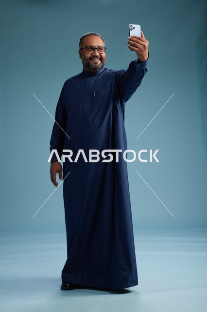 بورتريه لرجل عربي  سعودي  خليجي بثوب أزرق يلتقط سيلفي باستخدام الهاتف المحمول, ايماءات السعاده والضحك, اشارات وحركات متنوعه , الجسم بالطول الكامل, خلفيه زرقاء