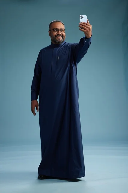 A portrait of an Arab Saudi Gulf man in a blue thobe taking a selfie with a mobile phone, expressions of happiness and laughter, various gestures and movements, full-body, blue background.