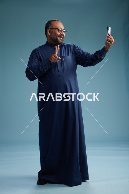 بورتريه لرجل عربي  سعودي  خليجي بثوب أزرق يلتقط سيلفي باستخدام الهاتف المحمول, ايماءات السعاده والضحك, اشارات وحركات متنوعه , الجسم بالطول الكامل, خلفيه زرقاء