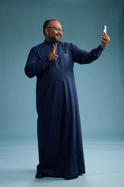 A portrait of an Arab Saudi Gulf man in a blue thobe taking a selfie with a mobile phone, expressions of happiness and laughter, various gestures and movements, full-body, blue background.