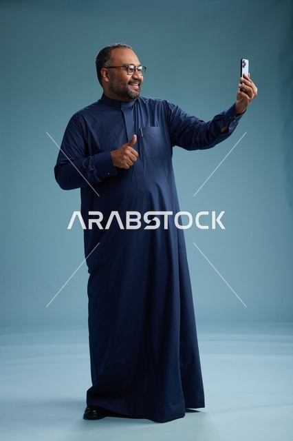 بورتريه لرجل عربي  سعودي  خليجي بثوب أزرق يلتقط سيلفي باستخدام الهاتف المحمول, ايماءات السعاده والضحك, اشارات وحركات متنوعه , الجسم بالطول الكامل, خلفيه زرقاء