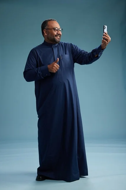 A portrait of an Arab Saudi Gulf man in a blue thobe taking a selfie with a mobile phone, expressions of happiness and laughter, various gestures and movements, full-body, blue background.