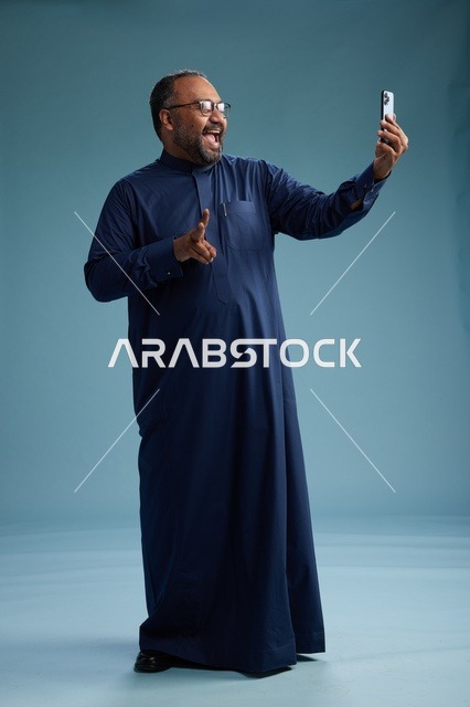 بورتريه لرجل عربي  سعودي  خليجي بثوب أزرق يلتقط سيلفي باستخدام الهاتف المحمول, ايماءات السعاده والضحك, اشارات وحركات متنوعه , الجسم بالطول الكامل, خلفيه زرقاء