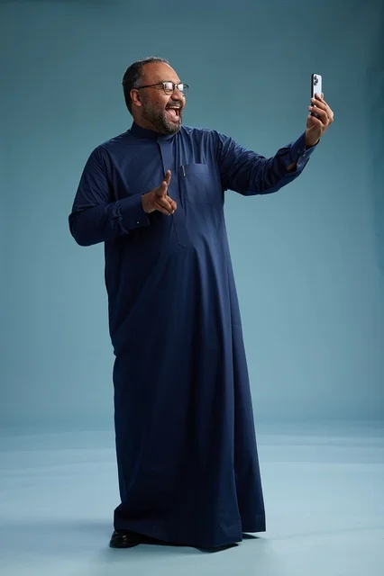 بورتريه لرجل عربي  سعودي  خليجي بثوب أزرق يلتقط سيلفي باستخدام الهاتف المحمول, ايماءات السعاده والضحك, اشارات وحركات متنوعه , الجسم بالطول الكامل, خلفيه زرقاء