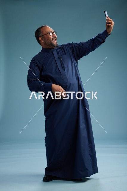 بورتريه لرجل عربي  سعودي  خليجي بثوب أزرق يلتقط سيلفي باستخدام الهاتف المحمول, ايماءات السعاده والضحك, اشارات وحركات متنوعه , الجسم بالطول الكامل, خلفيه زرقاء