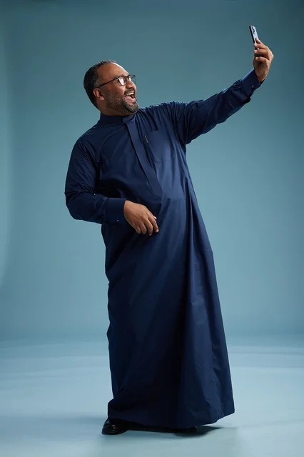 A portrait of an Arab Saudi Gulf man in a blue thobe taking a selfie with a mobile phone, expressions of happiness and laughter, various gestures and movements, full-body, blue background.