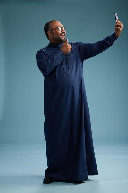 A portrait of an Arab Saudi Gulf man in a blue thobe taking a selfie with a mobile phone, expressions of happiness and laughter, various gestures and movements, full-body, blue background.