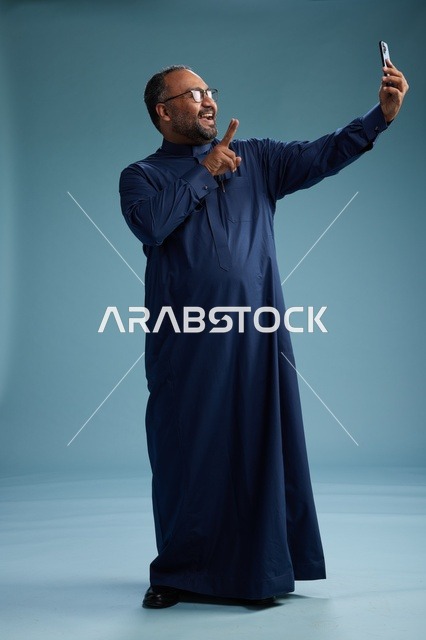 بورتريه لرجل عربي  سعودي  خليجي بثوب أزرق يلتقط سيلفي باستخدام الهاتف المحمول, ايماءات السعاده والضحك, اشارات وحركات متنوعه , الجسم بالطول الكامل, خلفيه زرقاء