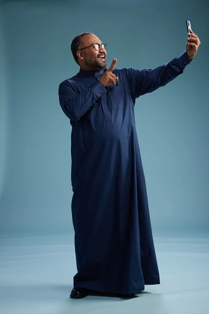 A portrait of an Arab Saudi Gulf man in a blue thobe taking a selfie with a mobile phone, expressions of happiness and laughter, various gestures and movements, full-body, blue background.