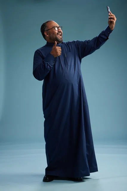 A portrait of an Arab Saudi Gulf man in a blue thobe taking a selfie with a mobile phone, expressions of happiness and laughter, various gestures and movements, full-body, blue background.