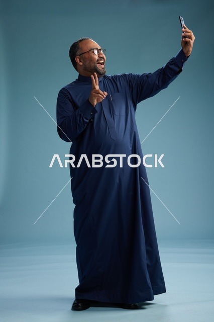 بورتريه لرجل عربي  سعودي  خليجي بثوب أزرق يلتقط سيلفي باستخدام الهاتف المحمول, ايماءات السعاده والضحك, اشارات وحركات متنوعه , الجسم بالطول الكامل, خلفيه زرقاء