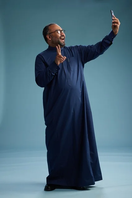 A portrait of an Arab Saudi Gulf man in a blue thobe taking a selfie with a mobile phone, expressions of happiness and laughter, various gestures and movements, full-body, blue background.