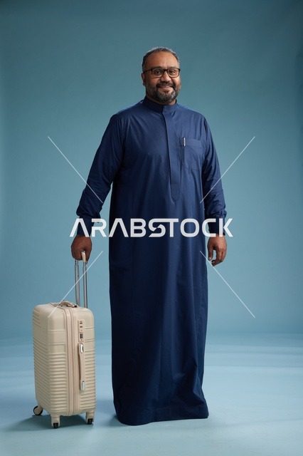 بورتيه لرجل  عربي سعودي  خليجي بثوب أزرق واقف مع حقيبة سفر, الاستعداد للسفر  والسياحه, الجسم بالطول الكامل, خلفيه زرقاء