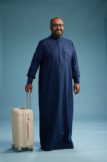 A portrait of a Saudi Arabian Gulf man in a blue thobe standing with a suitcase, ready for travel and tourism, full-body shot, blue background.