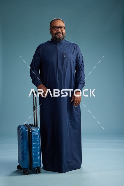 بورتيه لرجل  عربي سعودي  خليجي بثوب أزرق واقف مع حقيبة سفر, الاستعداد للسفر  والسياحه, الجسم بالطول الكامل, خلفيه زرقاء