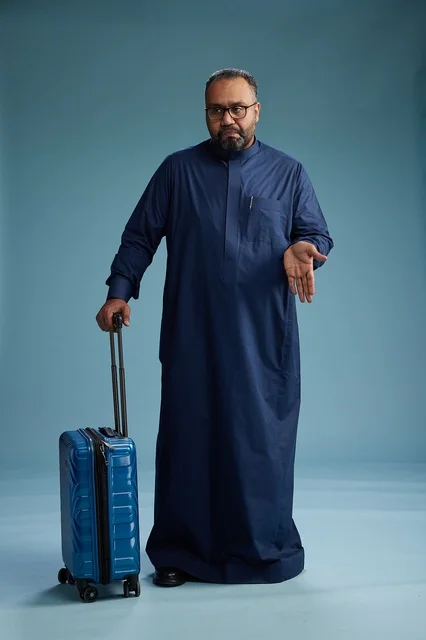 A portrait of a Saudi Arabian Gulf man in a blue thobe standing with a suitcase, ready for travel and tourism, full-body shot, blue background.