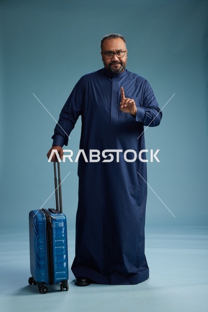 A portrait of a Saudi Arabian Gulf man in a blue thobe standing with a suitcase, ready for travel and tourism, full-body shot, blue background.
