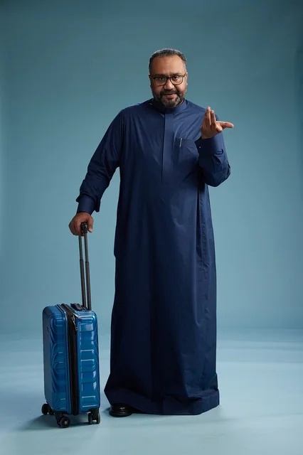 A portrait of a Saudi Arabian Gulf man in a blue thobe standing with a suitcase, ready for travel and tourism, full-body shot, blue background.