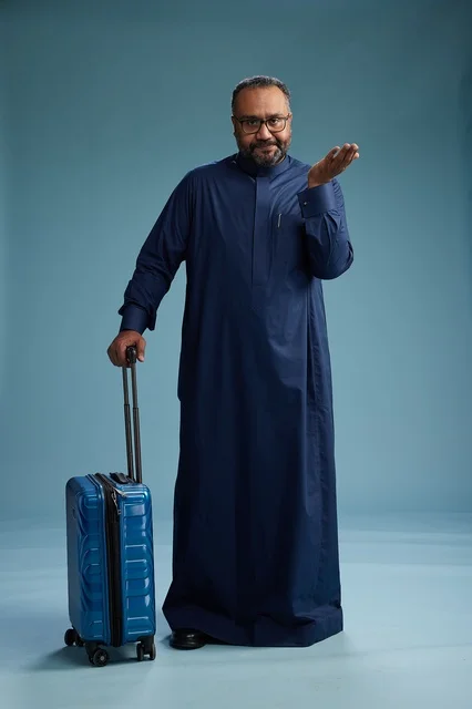 A portrait of a Saudi Arabian Gulf man in a blue thobe standing with a suitcase, ready for travel and tourism, full-body shot, blue background.