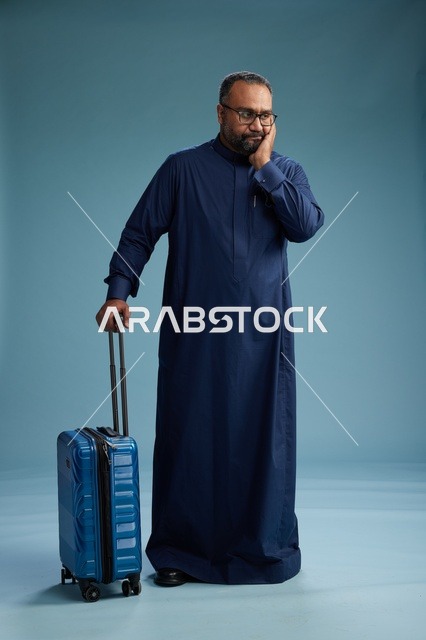 بورتيه لرجل  عربي سعودي  خليجي بثوب أزرق واقف مع حقيبة سفر, الاستعداد للسفر  والسياحه, الجسم بالطول الكامل, خلفيه زرقاء