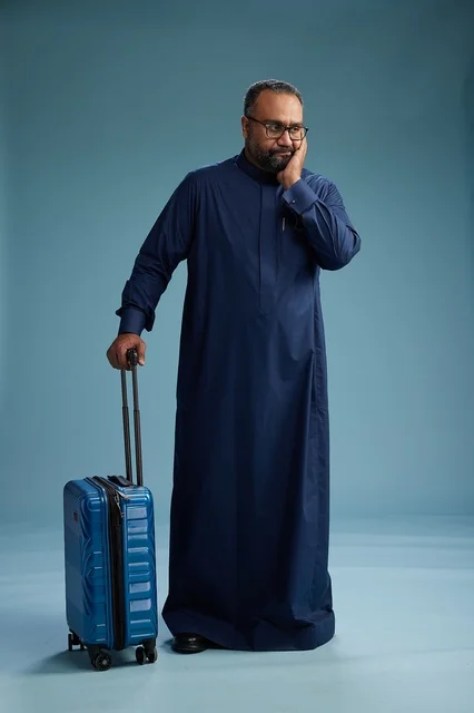 A portrait of a Saudi Arabian Gulf man in a blue thobe standing with a suitcase, ready for travel and tourism, full-body shot, blue background.
