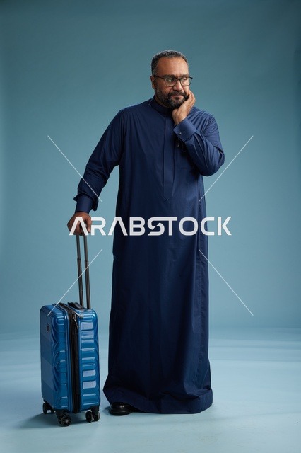 بورتيه لرجل  عربي سعودي  خليجي بثوب أزرق واقف مع حقيبة سفر, الاستعداد للسفر  والسياحه, الجسم بالطول الكامل, خلفيه زرقاء